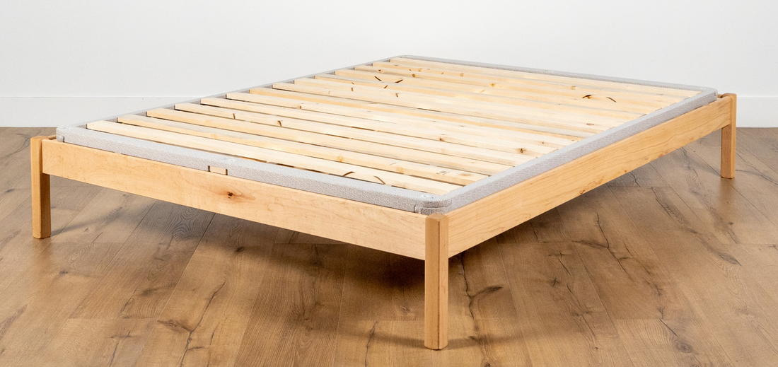 Wood slat bunkie board resting on a low platform bed frame, with a fabric-wrapped edge and natural wood legs visible